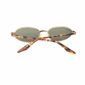 Puede incluir: Gafas de sol ovaladas con montura dorada y lentes oscuras. Las patillas presentan un dise&ntilde;o de carey. Las gafas de sol est&aacute;n sobre un fondo blanco.
