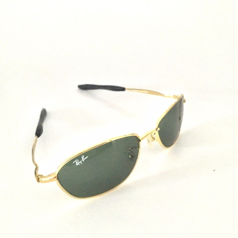 May include: Gold-tone Ray-Ban sunglasses with green lenses. The sunglasses have a thin, rectangular frame.