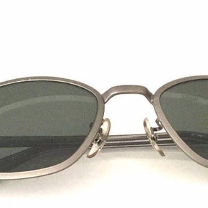 May include: Silver-toned metal sunglasses with oval-shaped lenses and a dark gray tint. The sunglasses have a thin metal frame and a Ray-Ban logo on the left lens.