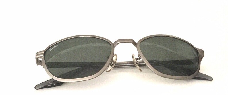 May include: Silver-toned metal sunglasses with oval-shaped lenses and a dark gray tint. The sunglasses have a thin metal frame and a Ray-Ban logo on the left lens.