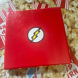 May include: A red square canvas with a yellow lightning bolt inside a white circle. The lightning bolt is the logo for the Flash, a superhero.