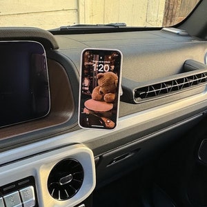 May include: A white phone mount attached to the dashboard of a car. The phone is displaying a teddy bear on the screen with the time 1:20.