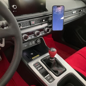 May include: Car interior showing a smartphone mounted on the dashboard, displaying the time 2:36 on a blue screen. The dashboard has a black infotainment screen, silver trim, and red interior. The gear stick has a red knob.