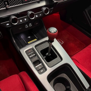 May include: Interior view of a car's console featuring a red and silver gear shift knob. The console has a silver trim, black accents, and red carpeting. A phone mount is visible on the dashboard.
