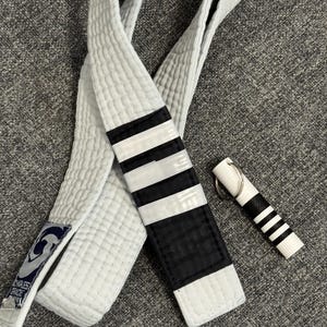 Peut inclure: Une ceinture d'arts martiaux blanche avec une extrémité noire ornée de trois bandes blanches et un petit écusson rectangulaire portant le texte Charles Gracie. À côté se trouve un porte-clés miniature blanc avec une section noire et trois bandes.
