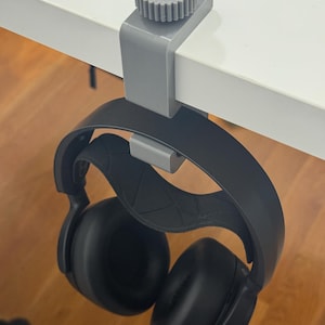 May include: A gray plastic headphone hanger with a circular, ridged tightening knob, clamped onto the edge of a white surface. A pair of black over-ear headphones with a textured, geometric patterned headband rests on the hook of the hanger.