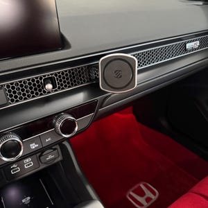 May include: A black and silver car phone mount fixed to a car dashboard. The mount is circular with a silver edge and a black centre. The dashboard features a honeycomb vent design and red floor mats.