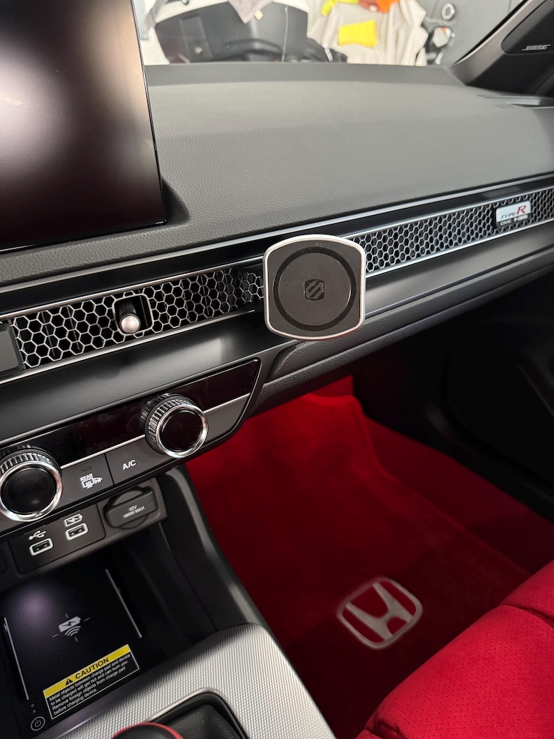 May include: A black and silver car phone mount fixed to a car dashboard. The mount is circular with a silver edge and a black centre. The dashboard features a honeycomb vent design and red floor mats.