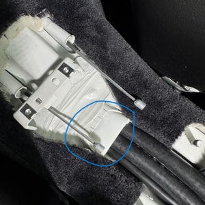 May include: A close-up of a car's interior showing a silver metal cable attached to a white plastic housing. The cable is secured with black tape and is surrounded by black wires.
