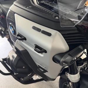 May include: Close-up of a motorcycle's front fairing. The fairing is silver and black, with the BMW logo visible. Features include black handles and a clear windshield. The motorcycle is parked indoors.