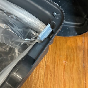 May include: Close-up of a black trash can with a plastic bag inside. The can has a silver latch that secures the bag in place.