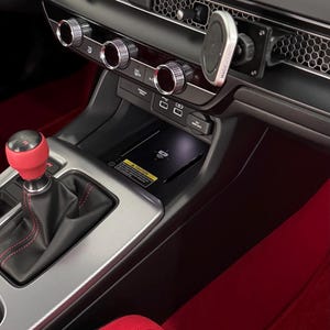 May include: Car interior showing the dashboard with a large screen, air vents, and control dials. The gear stick has a red knob. Red accents are visible on the seats and floor. The measurements are in centimeters.