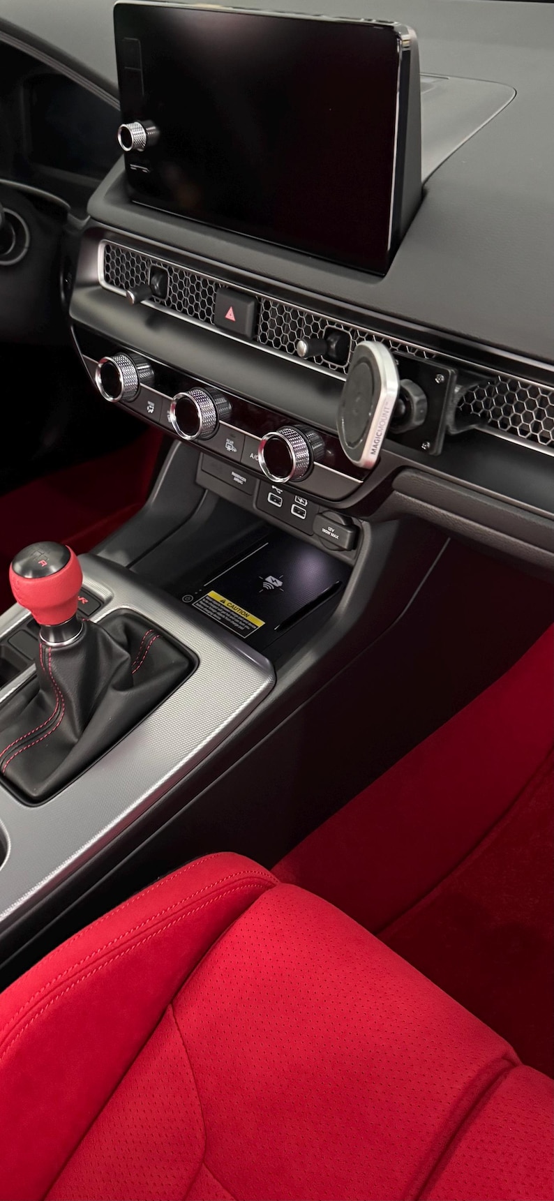 May include: Car interior showing the dashboard with a large screen, air vents, and control dials. The gear stick has a red knob. Red accents are visible on the seats and floor. The measurements are in centimeters.