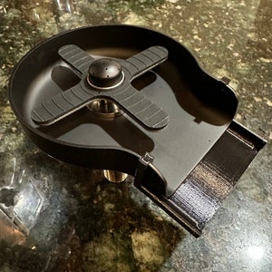 May include: A black matte cup rinser featuring a circular basin with a central cross-shaped sprayer arm. The device includes a drainage spout extending from the side and a threaded metal pipe connector on the underside for installation.