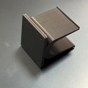 May include: A dark brown, 3D-printed bracket with a rectangular base and a curved, U-shaped support. The base has four small holes. The bracket is designed to hold an object. The item is made of plastic.