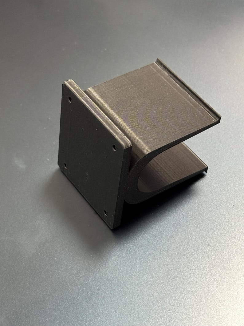 May include: A dark brown, 3D-printed bracket with a rectangular base and a curved, U-shaped support. The base has four small holes. The bracket is designed to hold an object. The item is made of plastic.