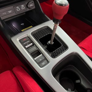 May include: Interior car detail featuring a red leather gear shift knob with a silver top. The center console has a silver trim, black honeycomb pattern, and red leather seats. Buttons include "Drive Mode" and "Brake Hold".