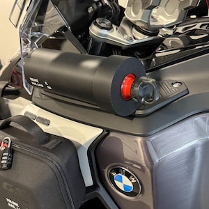 May include: A black cylindrical accessory with a red end cap is mounted on a motorcycle. The accessory has the text "VOLUME MAX. LOAD 1L 1kg". A black bag with a combination lock is also visible, along with the BMW logo.