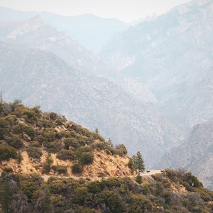 May include: A winding road through a mountainous landscape. The road is surrounded by trees and bushes, and the mountains in the distance are covered in a light haze.