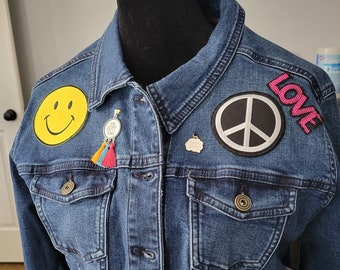 Peut inclure: Une veste en jean bleue avec des patchs, dont un smiley, un signe de paix et le mot "LOVE" en lettres roses. La veste a une fermeture à boutons et deux poches.