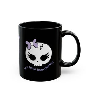 Puede incluir: Taza de cerámica negra con un diseño de calavera blanca, lazo morado y pestañas. El texto "you have been marked" está impreso en morado. La taza tiene un asa curva.