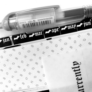 Peut inclure: Agenda en noir et blanc avec des onglets mensuels de janvier à juillet. Un stylo transparent est posé dessus. Les pages de l'agenda présentent un motif répété des lettres "H", "O" et "B". Le mot "currently" est imprimé sur les pages.
