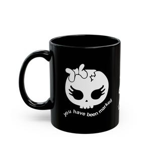 Puede incluir: Taza de cerámica negra con un diseño de calavera blanca. La calavera tiene formas de alas y una cabeza agrietada. El texto "you have been marked" está impreso en blanco. La taza tiene un asa curva.