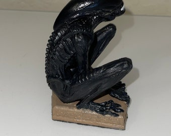 May include: A black Alien figurine, crouching on a rectangular base. The figurine has a smooth, glossy finish and detailed skeletal features. The base is a muted brown color.