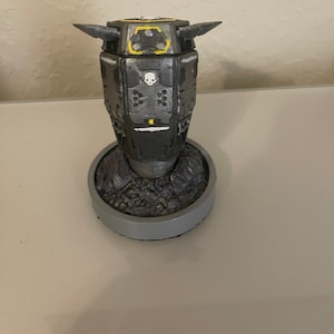 May include: A gray and black 3D printed model of a futuristic spaceship with yellow accents. The spaceship has a skull symbol on the front and is sitting on a gray base.