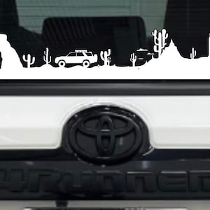 May include: White vinyl decal of a desert landscape with a jeep driving through it. The decal features a mountain range, cacti, and an archway. The decal is on the back window of a white car.