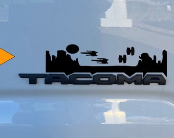 May include: A white Toyota Tacoma truck with a black "TACOMA" emblem and a black decal of the Star Wars Millennium Falcon flying over a desert landscape.