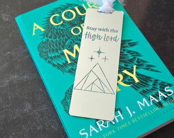 May include: A silver bookmark with a geometric mountain design and three gold stars. The bookmark has the text "Stay with the High Lord" engraved on it.