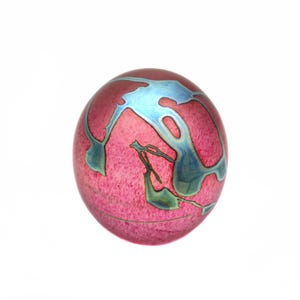 May include: A spherical glass paperweight featuring a vibrant magenta pink base with a textured, mottled appearance. The surface is adorned with swirling, iridescent blue and teal metallic patterns that create a fluid, organic design across the sphere.