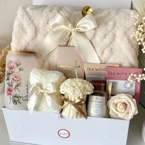 May include: A white gift box filled with self-care items. Includes a fluffy blanket, a floral-decorated glass, a small towel with a bow, a candle, bath salts, tea, and a rose-shaped soap. The box is tied with a large white bow.