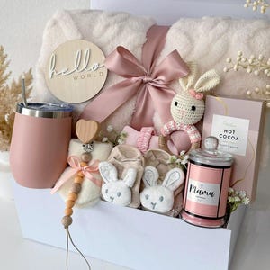 May include: A gift set in a white box containing a cream-colored plush blanket with a pink ribbon, a pink tumbler, a wooden pacifier clip with a heart, a crochet bunny rattle, beige baby booties with bunny faces, and a candle labeled Mama.