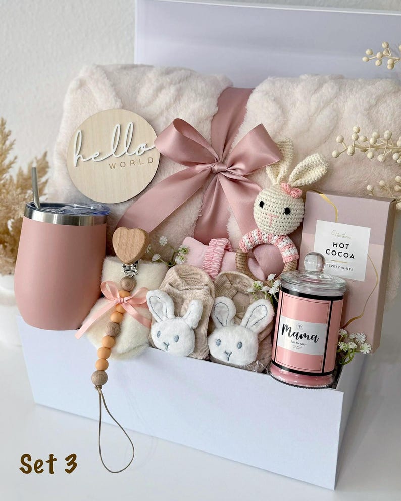 May include: A gift set in a white box containing a cream-colored plush blanket with a pink ribbon, a pink tumbler, a wooden pacifier clip with a heart, a crochet bunny rattle, beige baby booties with bunny faces, and a candle labeled Mama.