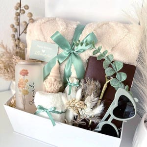May include: A white gift box labeled "Set 1" filled with a beige blanket tied with a mint green ribbon, a candle with floral design, a small gnome candle, a notebook, sunglasses, and a card that says "Happy Birthday! Beautiful."