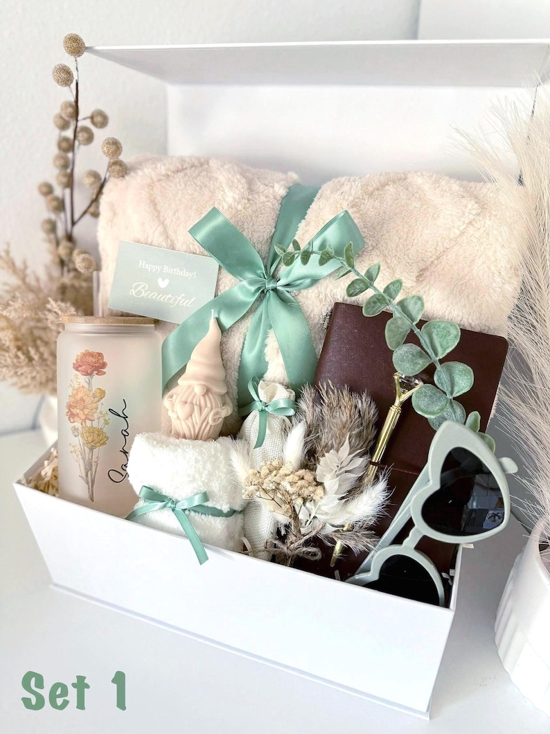 May include: A white gift box labeled "Set 1" filled with a beige blanket tied with a mint green ribbon, a candle with floral design, a small gnome candle, a notebook, sunglasses, and a card that says "Happy Birthday! Beautiful."
