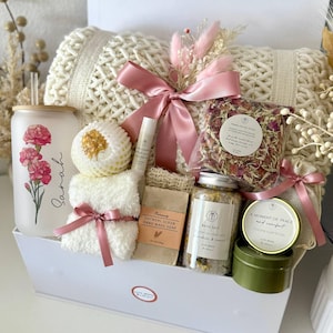May include: A white gift box containing self-care products. It features a cream-coloured knitted blanket, a glass tumbler with a floral design, oatmeal soap, bath salts, a small candle, and a pink ribbon. The text "Set 6" is at the bottom.
