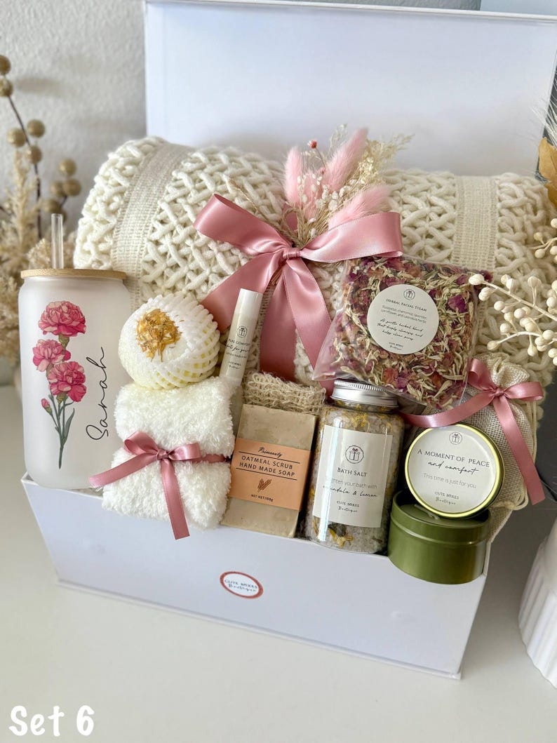 May include: A white gift box containing self-care products. It features a cream-coloured knitted blanket, a glass tumbler with a floral design, oatmeal soap, bath salts, a small candle, and a pink ribbon. The text "Set 6" is at the bottom.