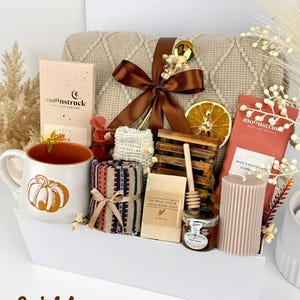 May include: A gift set containing a beige knit blanket with a brown ribbon, a white mug with an orange pumpkin, two Moonstruck chocolate bars, a patterned cloth, oatmeal soap, a wooden honey dipper, a jar of honey, and a ribbed candle.