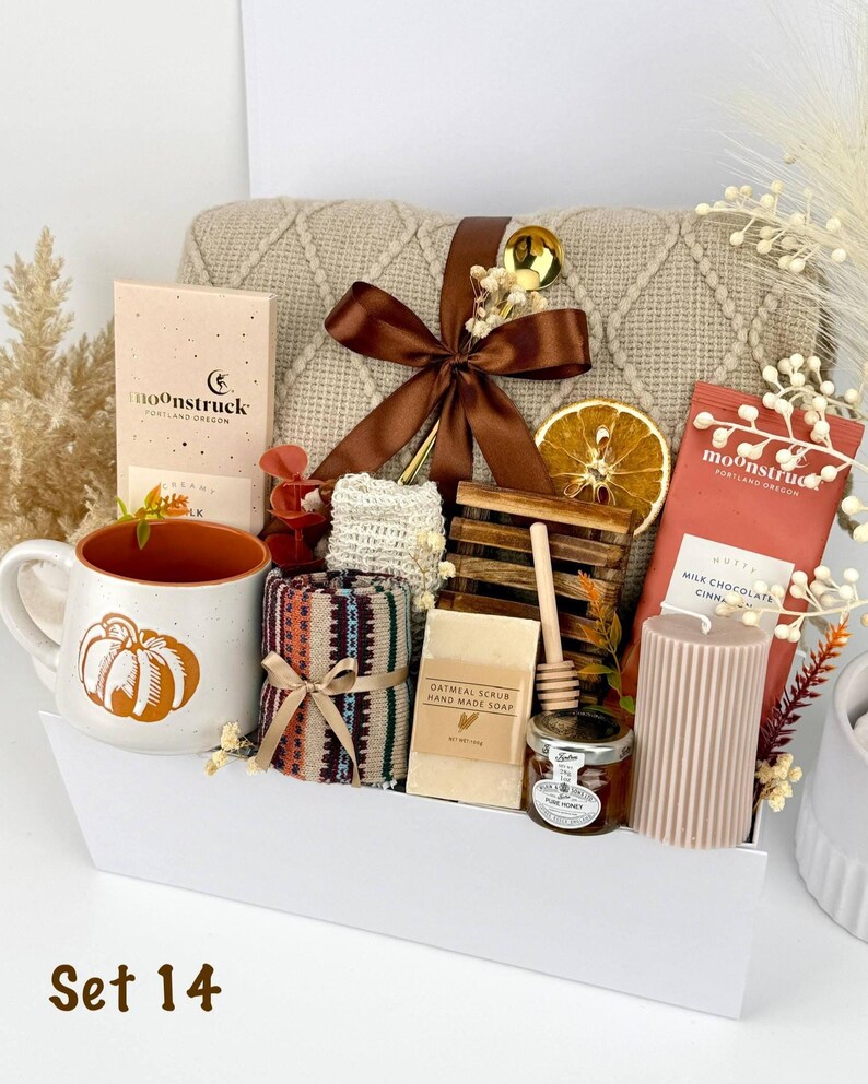 May include: A gift set containing a beige knit blanket with a brown ribbon, a white mug with an orange pumpkin, two Moonstruck chocolate bars, a patterned cloth, oatmeal soap, a wooden honey dipper, a jar of honey, and a ribbed candle.