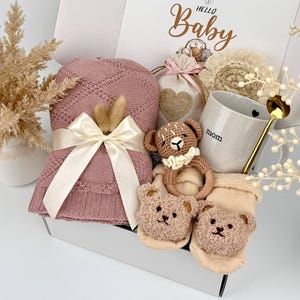 May include: A gift box labeled Hello Baby containing a dusty pink knitted blanket with a cream ribbon, a beige drawstring bag with a heart, a speckled mug with the text mom, a crochet bear rattle, and two beige baby socks with bear faces.