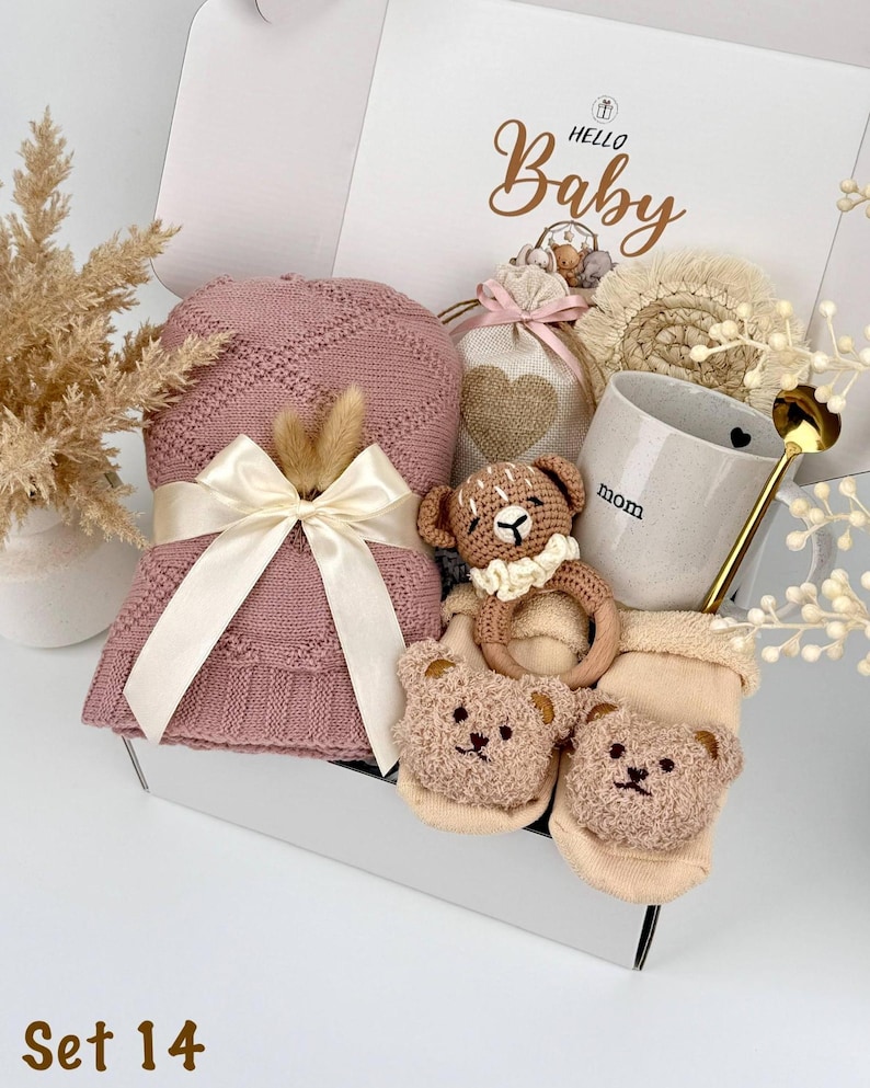 May include: A gift box labeled Hello Baby containing a dusty pink knitted blanket with a cream ribbon, a beige drawstring bag with a heart, a speckled mug with the text mom, a crochet bear rattle, and two beige baby socks with bear faces.