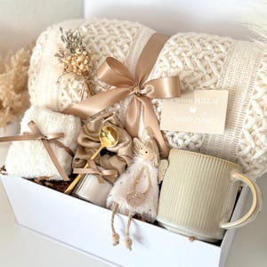 May include: A gift set containing a cream-colored cable knit throw blanket tied with a tan ribbon, a small white fuzzy cloth, a tan scrunchie, a gold spoon, a small angel figurine, and a ribbed ceramic mug with the text A season FULL of Simple Joys.