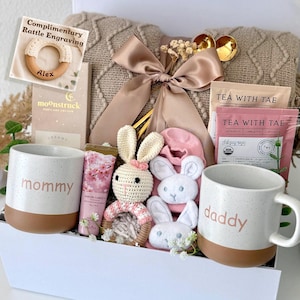May include: A gift set containing a beige cable-knit blanket with a satin bow, two speckled ceramic mugs labeled mommy and daddy, a crochet bunny rattle, pink baby socks, a tube of Sakura Garden hand cream, and Tea With Tae tea bags.