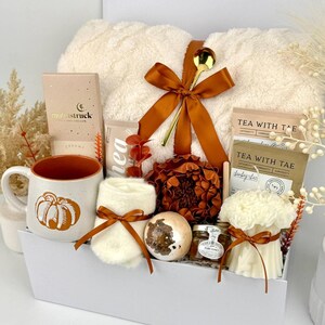 May include: A gift set in a white box containing a cream-coloured fuzzy blanket tied with a brown ribbon and gold spoon, a speckled mug with a pumpkin graphic, a pair of cream socks, a bath bomb, a jar of honey, tea packets, and a floral candle.
