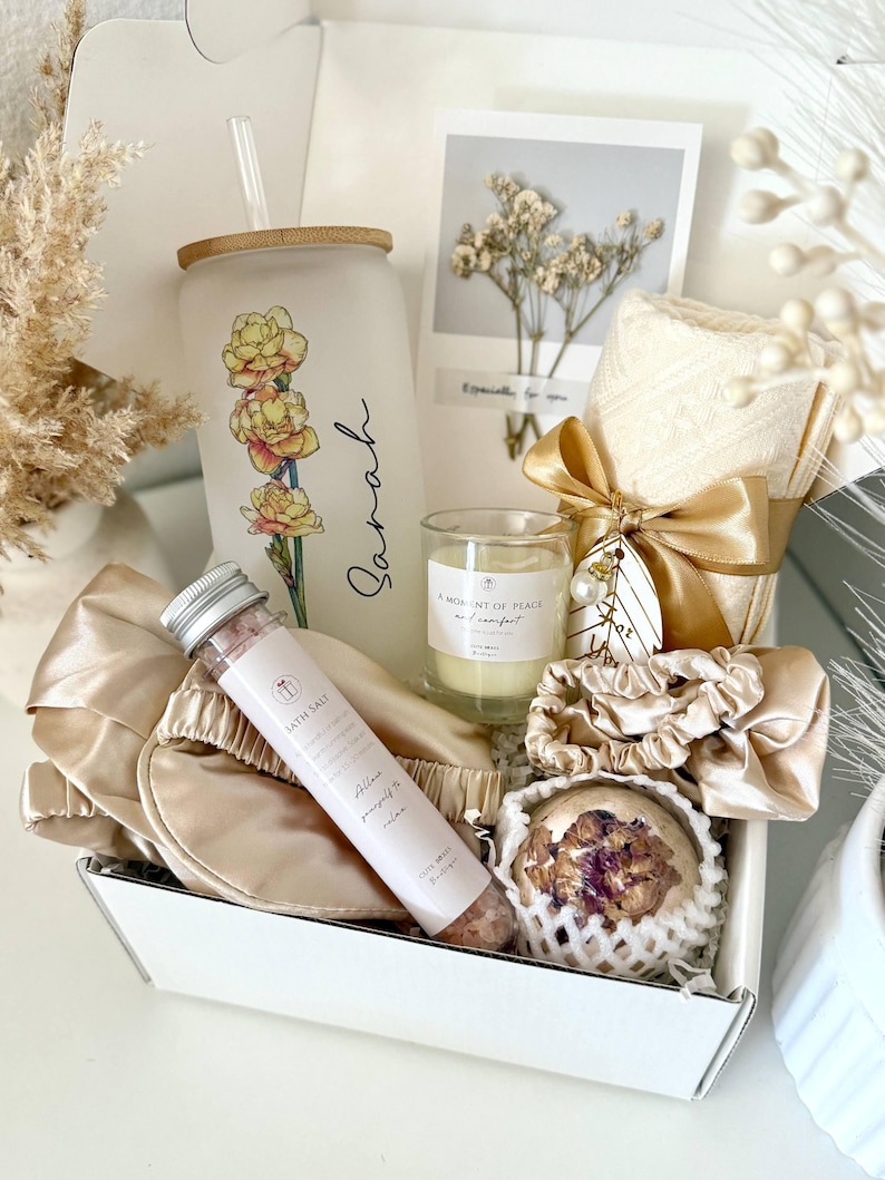 May include: A self-care gift box containing a glass tumbler with a floral design and the name "Sarah", a candle, bath salts, a sleep mask, a bath bomb, and a small card with dried flowers.