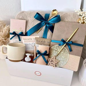 Puede incluir: Una cesta de regalo llena de una manta beige, chocolate, t&eacute;, miel, una taza y objetos decorativos. Los art&iacute;culos est&aacute;n atados con cintas color turquesa y presentados en una caja blanca. Cucharas doradas y un adorno en forma de estrella a&ntilde;aden un toque de elegancia.
