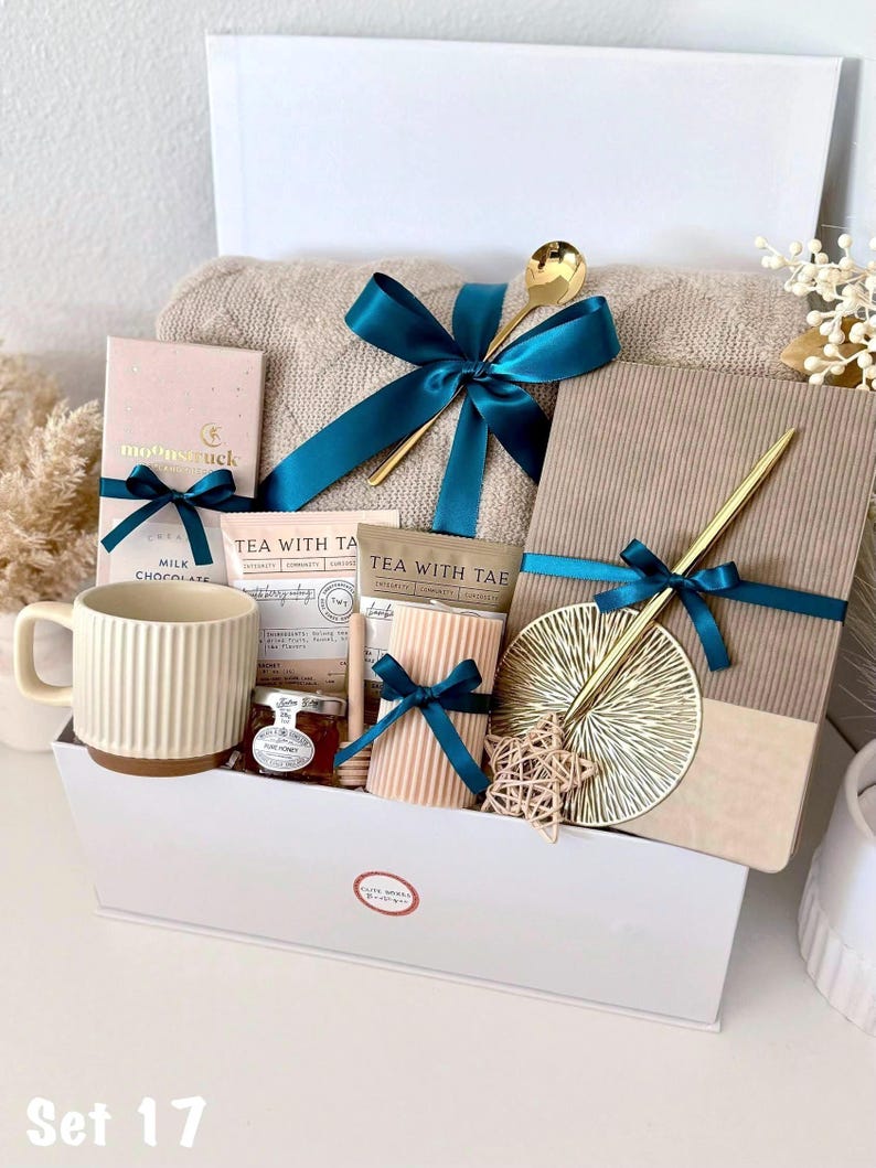 Puede incluir: Una cesta de regalo llena de una manta beige, chocolate, t&eacute;, miel, una taza y objetos decorativos. Los art&iacute;culos est&aacute;n atados con cintas color turquesa y presentados en una caja blanca. Cucharas doradas y un adorno en forma de estrella a&ntilde;aden un toque de elegancia.